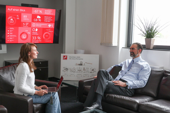 Eine Frau interviewt einen Mann in einem Büro, im Hintergrund werden auf einem Bildschirm Geschäftsdiagramme angezeigt. - © Fotos: Westfalen AG © Fotos: Westfalen AG