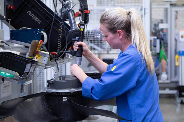 Eine Frau in blauer Arbeitskleidung montiert Maschinenteile an einem Industriearbeitsplatz in einer Fabrik. - © ebm-papst © ebm-papst