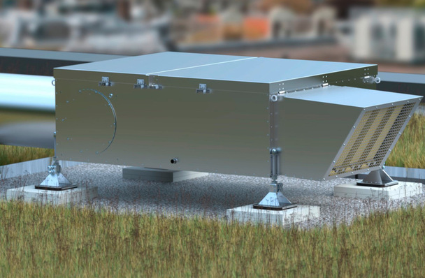 Ein großes Metalllüftungsgerät steht auf Stützen in einem grasbewachsenen Außenbereich, während die Gebäude im Hintergrund verschwimmen. - © Bild: Aereco