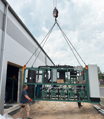 Ein Kran hebt schwere Industrieanlagen außerhalb eines Gebäudes an, während zwei Arbeiter in der Nähe stehen. - © Bild: compact Kälttechnik © Bild: compact Kälttechnik