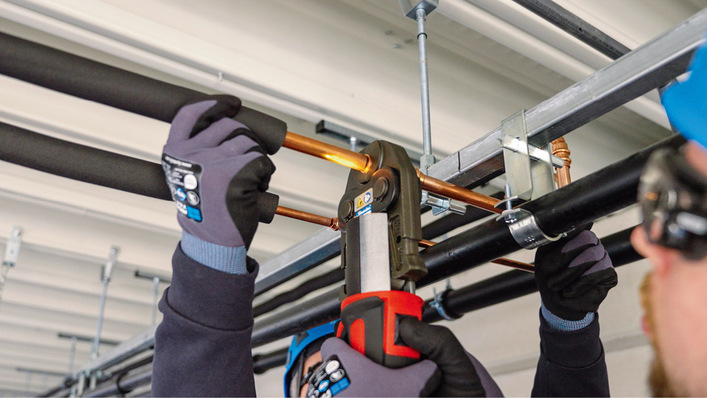 Ein Arbeiter verwendet ein Presswerkzeug, um Kupferrohre an der Decke zu verbinden. Er trägt Handschuhe und Schutzkleidung. - © Bild: Novopress