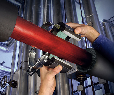 Hände, die in einer Industrieumgebung zwischen Metallrohren eine Isolierung an einem roten Rohr anbringen. - © Bild: Armacell © Bild: Armacell