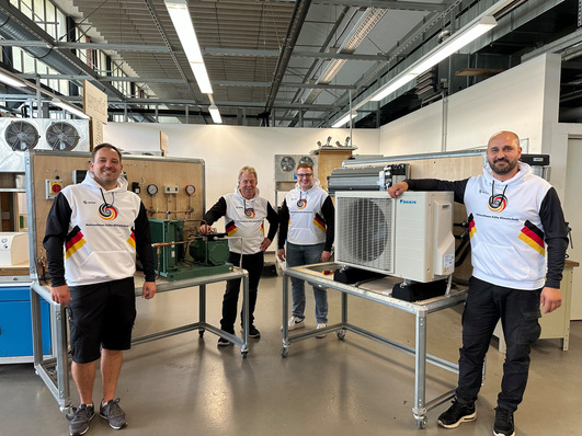 Vier Personen in passenden weißen Hemden stehen in einer Werkstatt mit HLK-Geräten und Werkzeugen und lächeln in die Kamera. - © KKE gGmbH, Duisburg) © KKE gGmbH, Duisburg)