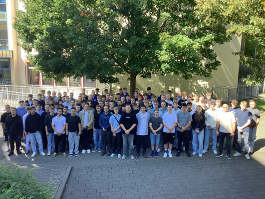 Eine große Gruppe von Menschen steht im Freien auf einer gepflasterten Fläche unter einem Baum zusammen und blickt in die Kamera. - © BSZ Vogtland © BSZ Vogtland